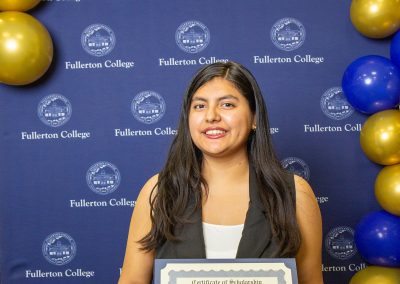A person smiles while holding a scholarship certificate in front of a blue backdrop with the Fullerton College logo and gold and blue balloons.