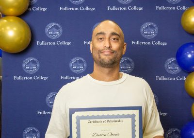 A person stands in front of a blue backdrop with gold and blue balloons, holding a scholarship certificate from Fullerton College.