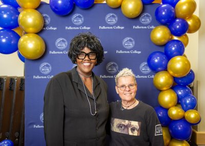 Two individuals pose together in front of a blue backdrop featuring the Fullerton College logo, framed by blue and gold balloons.