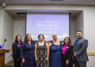 A group of six individuals poses for a photo in front of a presentation screen displaying "Theodore Grimm" and "Patricia A. Gallo Del Giorgio Osher" in a conference room setting.
