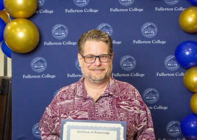 A person holds a scholarship certificate in front of a blue backdrop with the Fullerton College logo and gold and blue balloons.