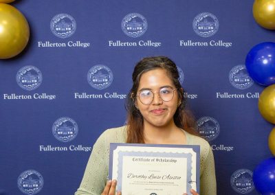 A person holds a scholarship certificate in front of a backdrop featuring the Fullerton College logo and blue and gold balloons.