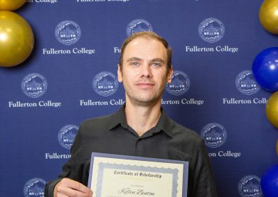A person stands in front of a blue backdrop with "Fullerton College" logos and blue and gold balloons, holding a certificate of scholarship.