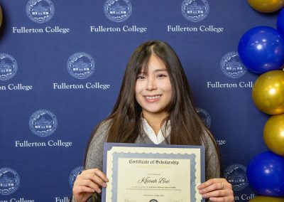 A person smiles while holding a scholarship certificate at a Fullerton College event, with blue and gold balloons decorating the background.