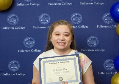 A person smiles while holding a scholarship certificate at a Fullerton College event, standing in front of a branded backdrop with balloons.