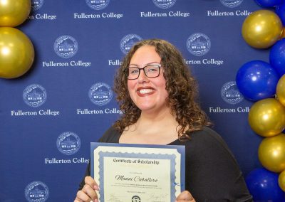 A person with curly hair and glasses smiles while holding a scholarship certificate in front of a blue backdrop adorned with gold and blue balloons and the Fullerton College logo.