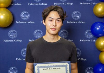 A person stands in front of a blue backdrop with gold and blue balloons, holding a certificate of scholarship from Fullerton College.