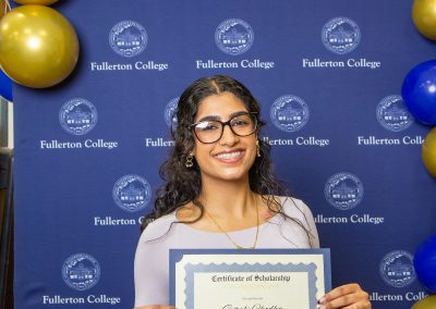 A person with dark hair and glasses smiles while holding a scholarship certificate in front of a blue backdrop with the Fullerton College logo and gold and blue balloons.