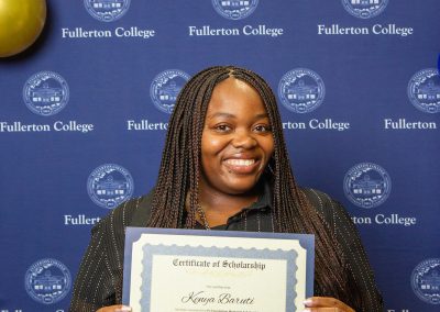 A person smiles while holding a scholarship certificate in front of a blue backdrop featuring the Fullerton College logo and balloons.