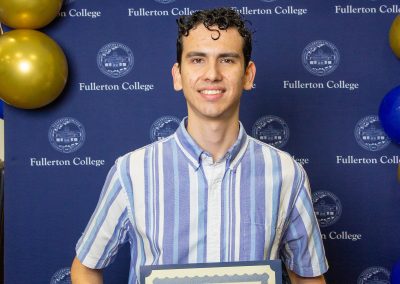 A young person smiles while holding a scholarship certificate in front of a blue backdrop with the Fullerton College logo and blue and gold balloons.