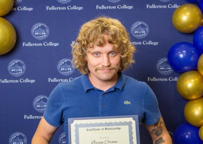 A person with curly hair and a mustache holds a scholarship certificate in front of a blue backdrop adorned with gold and blue balloons and the Fullerton College logo.