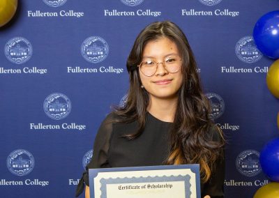A person with long dark hair and glasses stands in front of a blue backdrop with "Fullerton College" logos, holding a certificate of scholarship awarded on May 18, 2020.