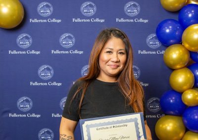 A person stands in front of a blue backdrop with "Fullerton College" logos and gold and blue balloons, holding a scholarship certificate.