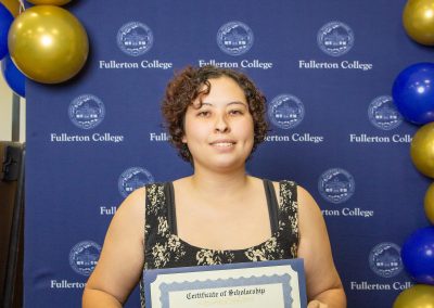 A person stands in front of a blue backdrop with gold and blue balloons, holding a certificate of scholarship from Fullerton College.