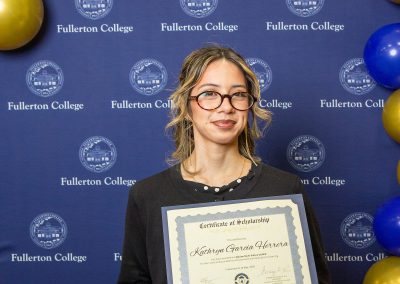A person with glasses holds a scholarship certificate in front of a blue backdrop featuring the Fullerton College logo and decorative balloons.