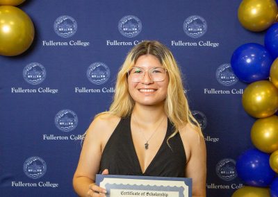 A person with blonde hair and glasses smiles while holding a scholarship certificate from Fullerton College, standing in front of a blue backdrop with gold and blue balloons.