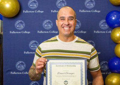 A person smiles while holding a scholarship certificate in front of a blue backdrop with the Fullerton College logo and gold and blue balloons.