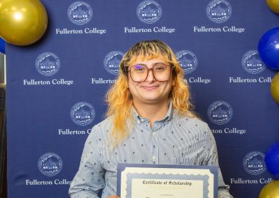 A person with long hair and glasses smiles while holding a scholarship certificate in front of a blue backdrop with the Fullerton College logo and gold and blue balloons.