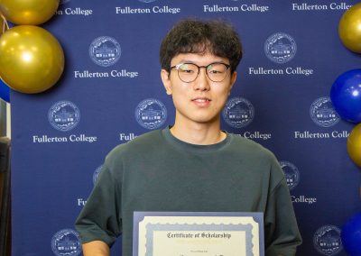 A young person holds a scholarship certificate in front of a blue backdrop with the Fullerton College logo and blue and gold balloons.