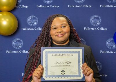 A person with long braided hair smiles while holding a scholarship certificate in front of a blue backdrop featuring the Fullerton College logo and gold and blue balloons.