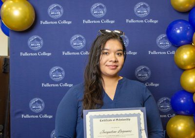 A person stands in front of a blue backdrop with gold and blue balloons, holding a scholarship certificate from Fullerton College.
