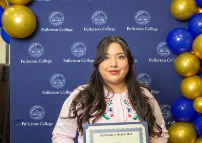 A person stands in front of a blue backdrop with gold and blue balloons, holding a certificate of scholarship from Fullerton College.