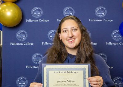 A person smiles while holding a scholarship certificate in front of a blue backdrop with the Fullerton College logo and gold and blue balloons.
