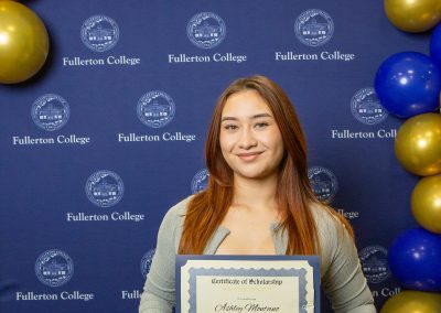 A person stands in front of a blue backdrop with the Fullerton College logo, holding a scholarship certificate, surrounded by blue and gold balloons.