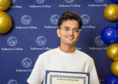 A person wearing glasses holds a scholarship certificate in front of a blue backdrop with "Fullerton College" logos and blue and gold balloons.