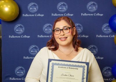 A person with glasses smiles while holding a scholarship certificate at a Fullerton College event, standing in front of a blue backdrop with gold and blue balloons.