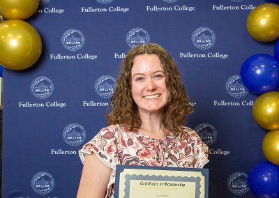 A person smiles while holding a scholarship certificate in front of a blue backdrop with the Fullerton College logo and gold and blue balloons.