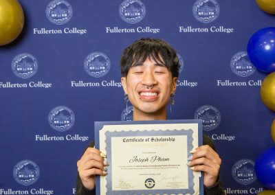 A person smiles broadly while holding a scholarship certificate in front of a blue backdrop with "Fullerton College" logos and blue and gold balloons.
