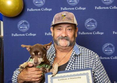 A person holds a small dog and a scholarship certificate in front of a Fullerton College backdrop adorned with balloons.