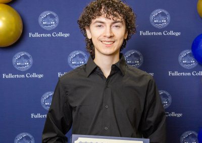 A person with curly hair stands in front of a blue backdrop featuring the Fullerton College logo and gold and blue balloons, holding a certificate of scholarship.