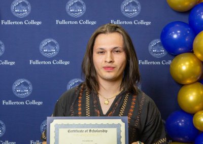 A person holds a scholarship certificate in front of a backdrop featuring the Fullerton College logo and blue and gold balloons.