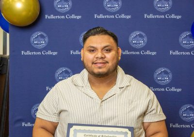 A person holds a scholarship certificate in front of a blue backdrop with the Fullerton College logo and gold and blue balloons.