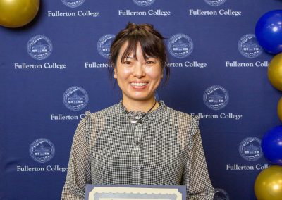 A person smiles while holding a scholarship certificate at a Fullerton College event, with blue and gold balloons decorating the background.