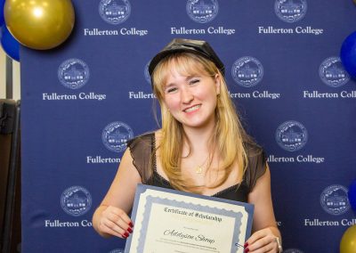 A person smiles while holding a scholarship certificate at a Fullerton College event, standing in front of a branded backdrop with blue and gold balloons.