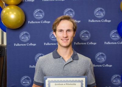 A person stands in front of a blue backdrop with gold and blue balloons, holding a certificate of scholarship from Fullerton College.