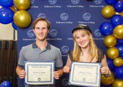 Two individuals stand in front of a blue backdrop adorned with gold and blue balloons, each holding a certificate of scholarship from Fullerton College.