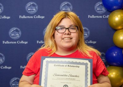 A person with blonde hair and glasses, wearing a red shirt, holds a scholarship certificate in front of a blue backdrop with the Fullerton College logo and gold and blue balloons.