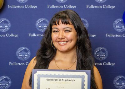 A person smiles while holding a scholarship certificate at a Fullerton College event, with balloons and a branded backdrop behind them.