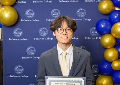 A person in formal attire holds a scholarship certificate in front of a blue backdrop with the Fullerton College logo and gold and blue balloons.