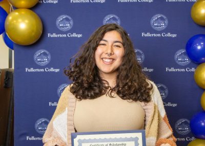 A person with long curly hair smiles while holding a scholarship certificate at a Fullerton College event, surrounded by blue and gold balloons.