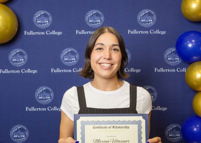 A person smiles while holding a scholarship certificate at a Fullerton College event, standing in front of a branded backdrop adorned with blue and gold balloons.