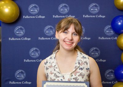 A person smiles while holding a scholarship certificate in front of a blue backdrop with the Fullerton College logo and gold and blue balloons.