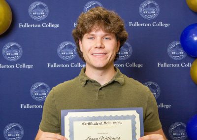 A young person holds a scholarship certificate in front of a blue backdrop featuring the Fullerton College logo and decorated with blue and gold balloons.