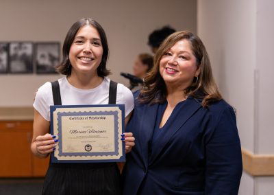 A young person holds a scholarship certificate while standing beside an adult, both smiling for the camera.