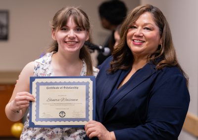 A young person holds a scholarship certificate while standing beside an adult, both smiling at the camera.