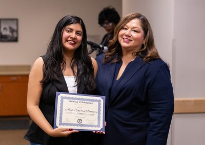 Two individuals stand together, one holding a framed certificate of scholarship, smiling for the camera in an indoor setting.